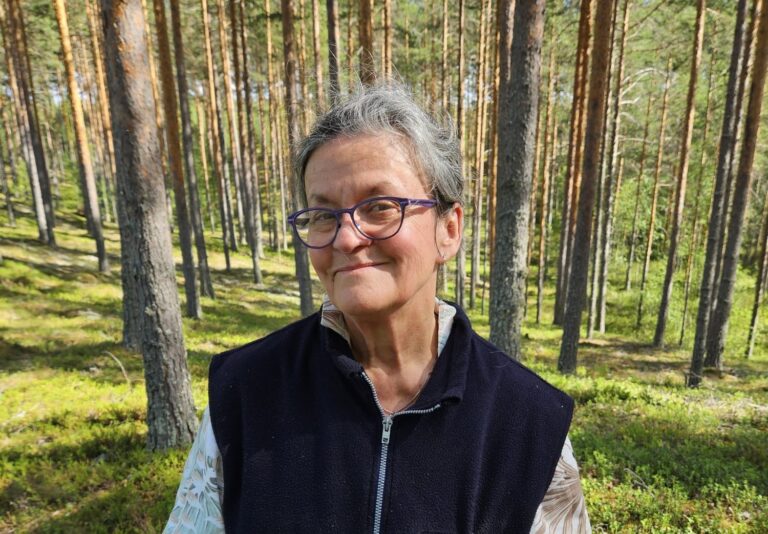 Nainen hymyilee mäntymetsässä.