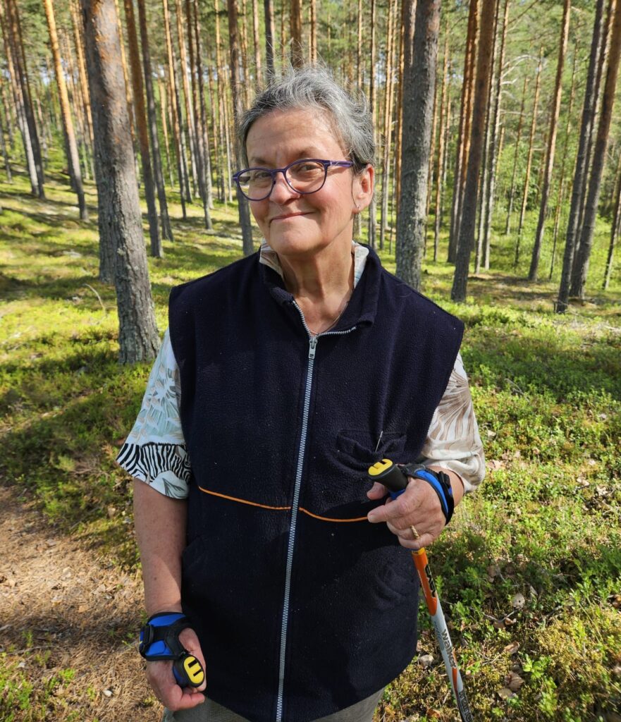 Nainen hymyilee mäntymetsässä sauvakävelyllä.